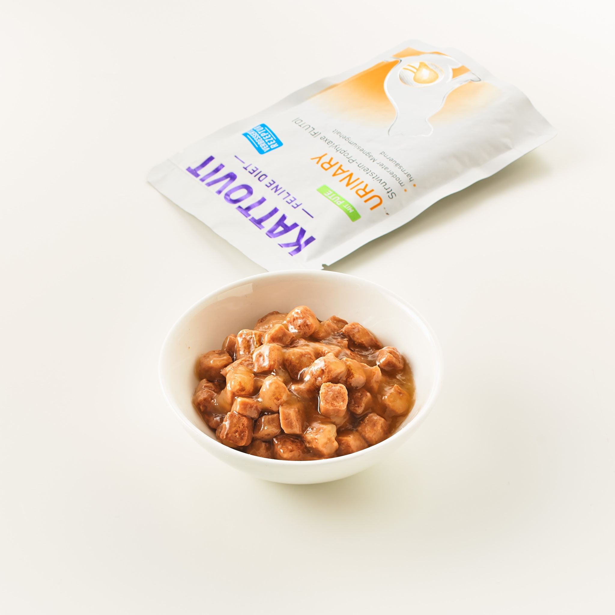 Eine Schüssel mit fleischigen Würfeln in Soße. Daneben liegt eine geöffnete Packung Katzenfutter KATTOVIT Urinary.