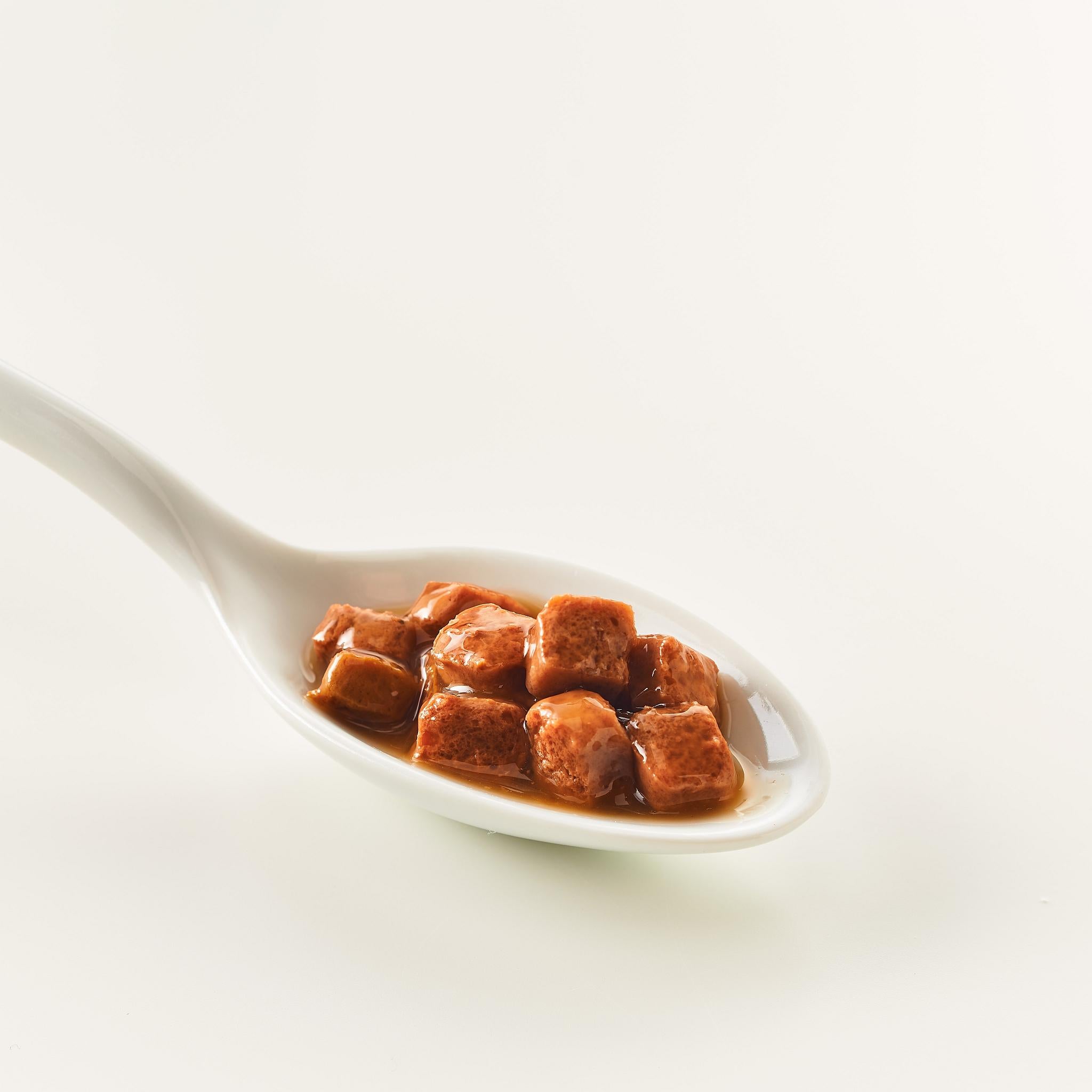 Weißer Löffel mit kleinen braunen Würfeln in Flüssigkeit auf weißem Hintergrund.