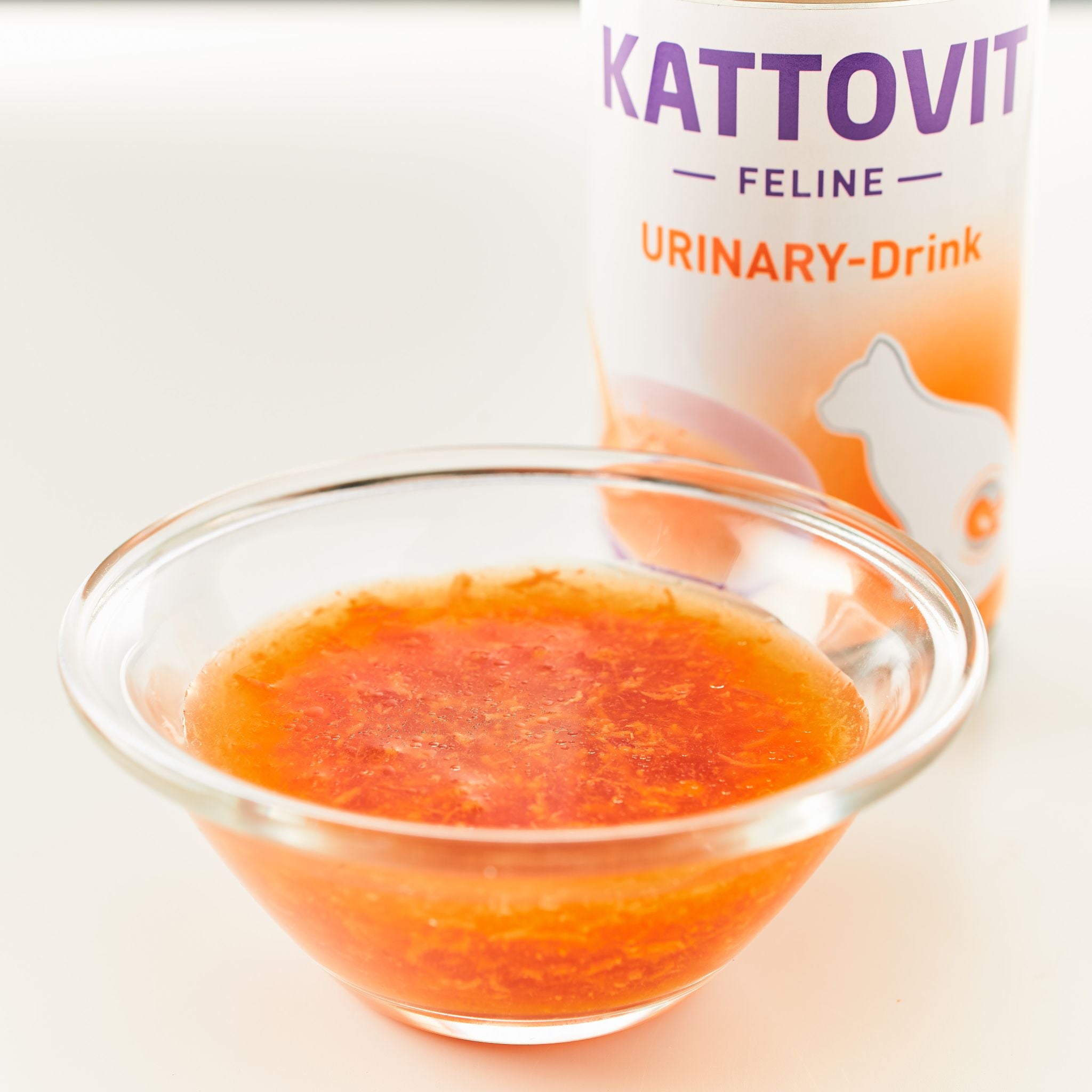 Eine kleine Glasschüssel mit einer orangefarbenen Flüssigkeit vor einer Verpackung eines Urinary-Drinks von KATTOVIT.