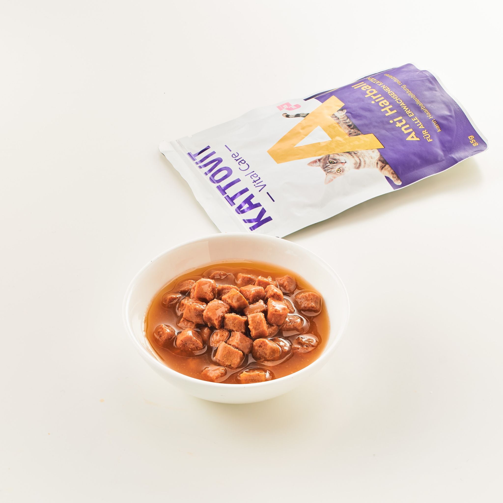 Eine weiße Schüssel mit braunen Fleischstückchen in Flüssigkeit. Daneben liegt eine aufgerissene Packung Katzenfutter mit der Aufschrift "KATTOVIT Anti Hairball".